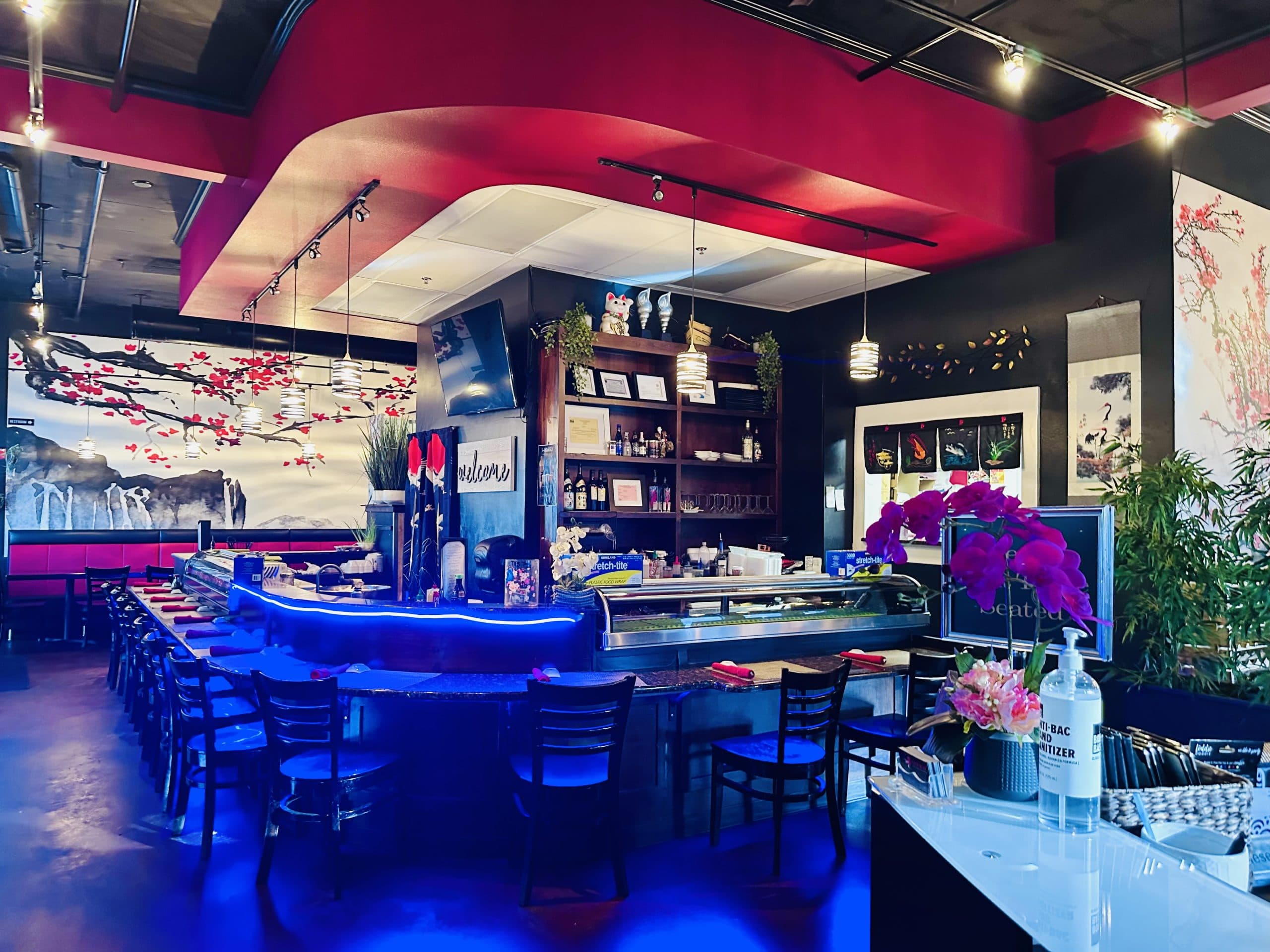 Genkai’s sushi bar, lit in blue, with the refrigerated fish case, cherry blossom murals along the wall and hanging scrolls